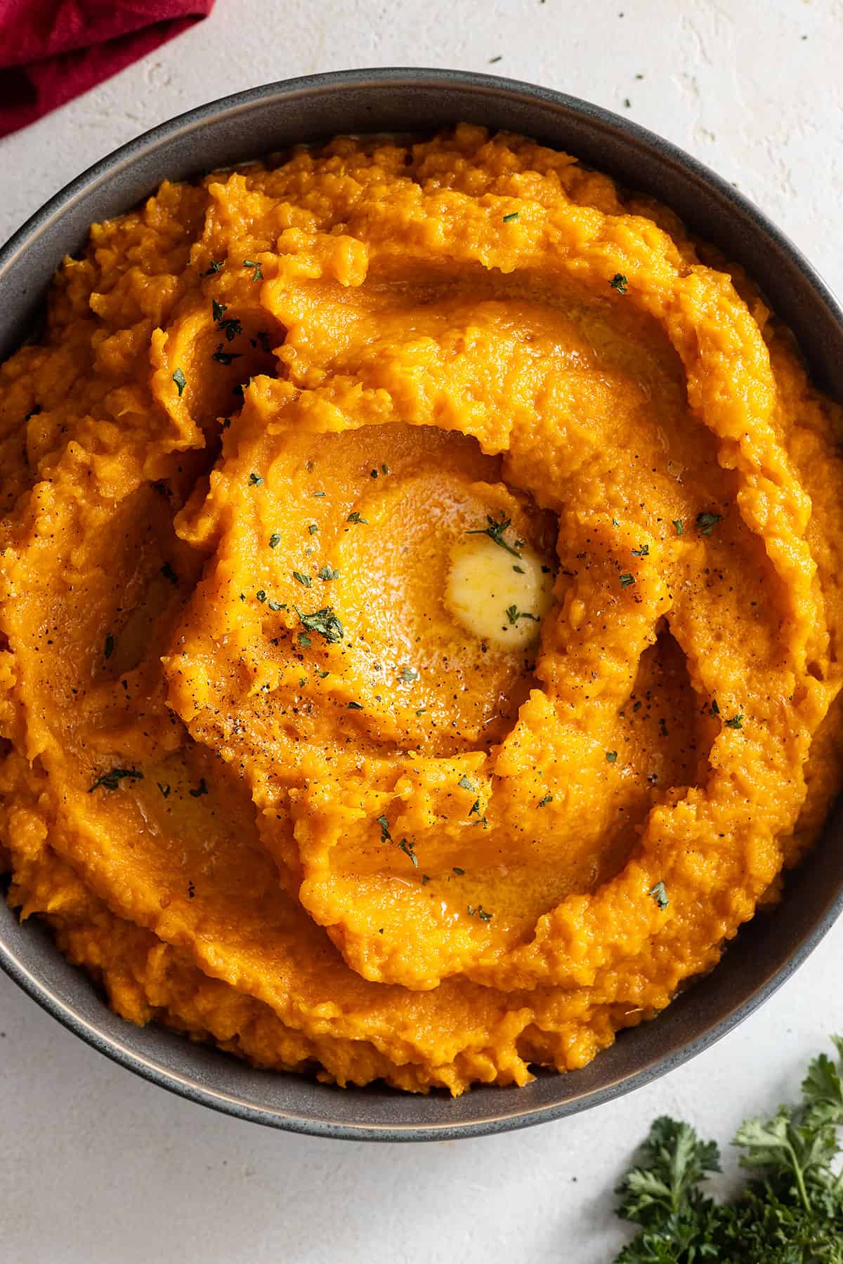 Overhead view of mashed sweet potatoes in a dark colored bowl. Garnished with black pepper, parsley, and melted butter. 
