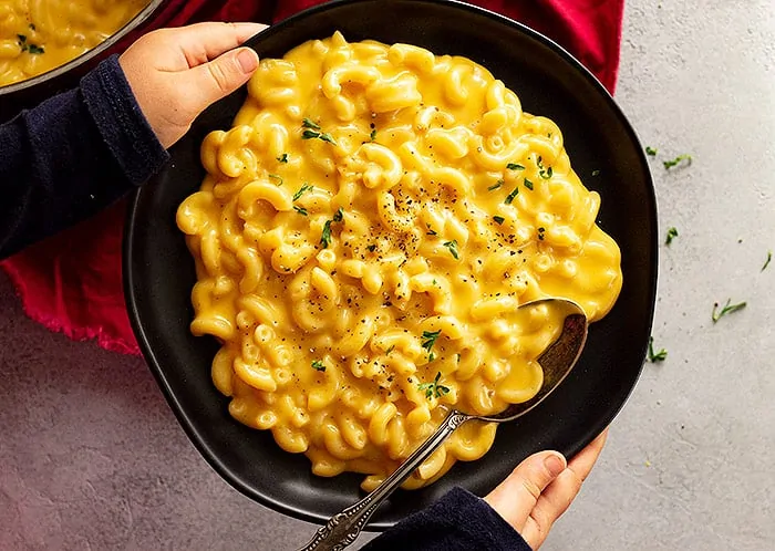 overhead: a child's hands holding a bowl of creamy stovetop mac and cheese garnished with black pepper and parsley