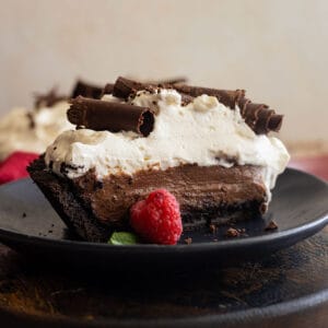 Slice of chocolate cream pie on a black plate. Garnished with chocolate curls and a fresh raspberry.