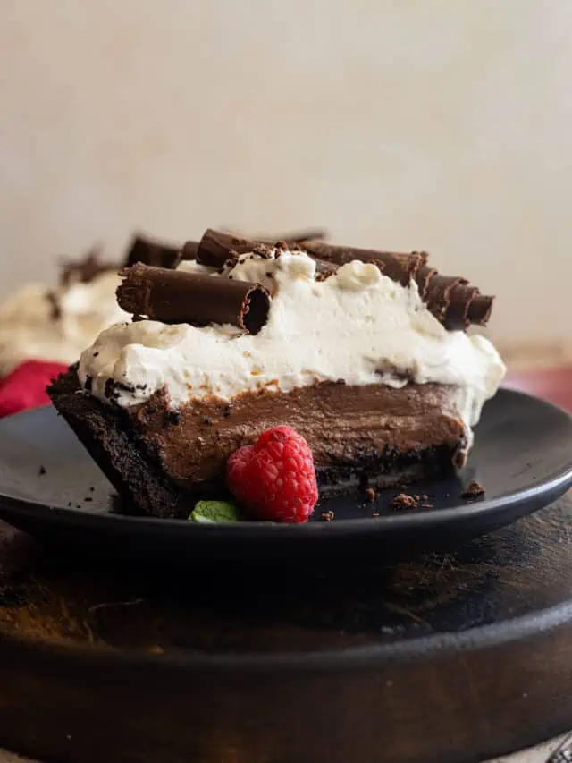 Slice of chocolate cream pie on a black plate. Garnished with chocolate curls and a fresh raspberry.