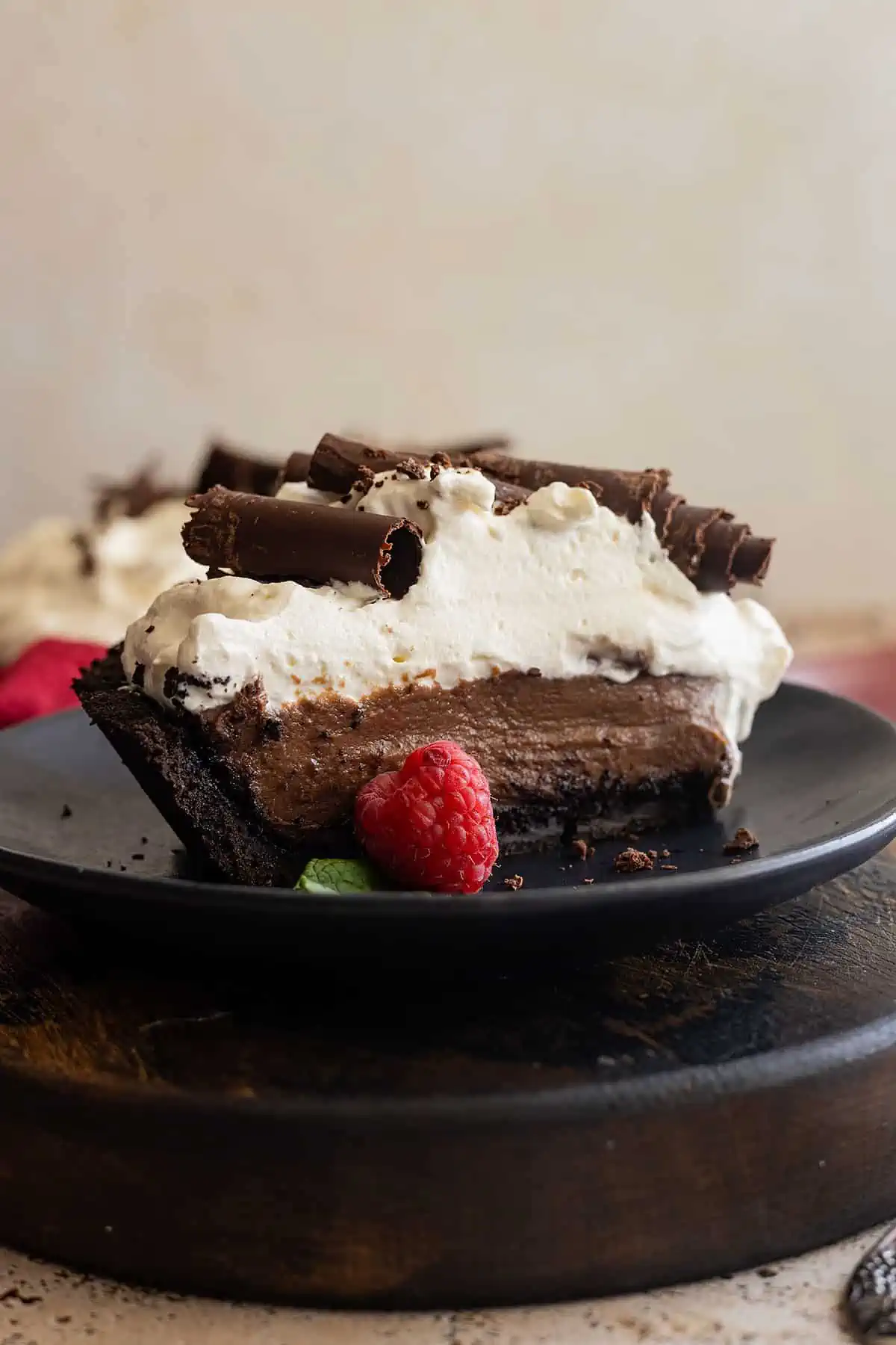 Slice of chocolate cream pie on a black plate. Garnished with chocolate curls and a fresh raspberry. 