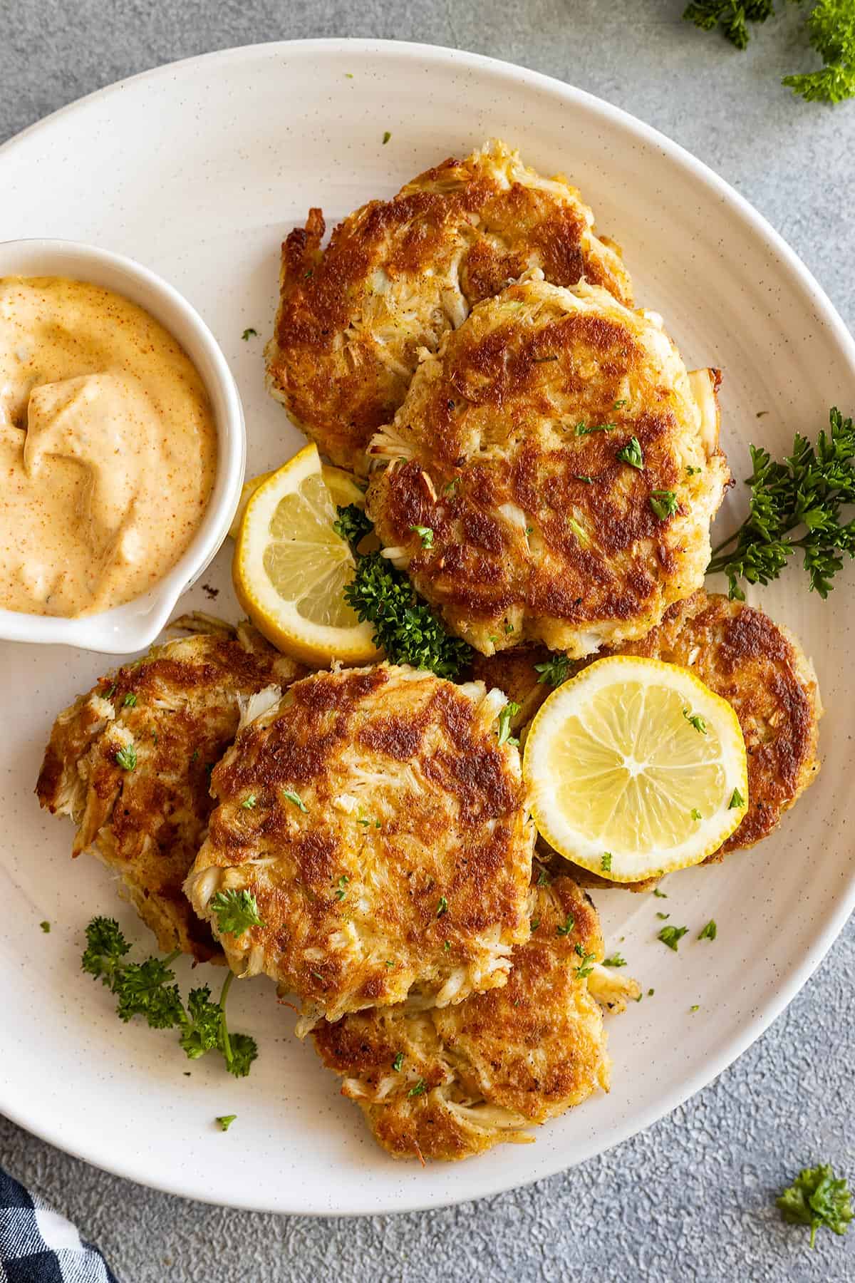 Overhead view of crab cakes on a white plate with dipping sauce and lemons for serving. 