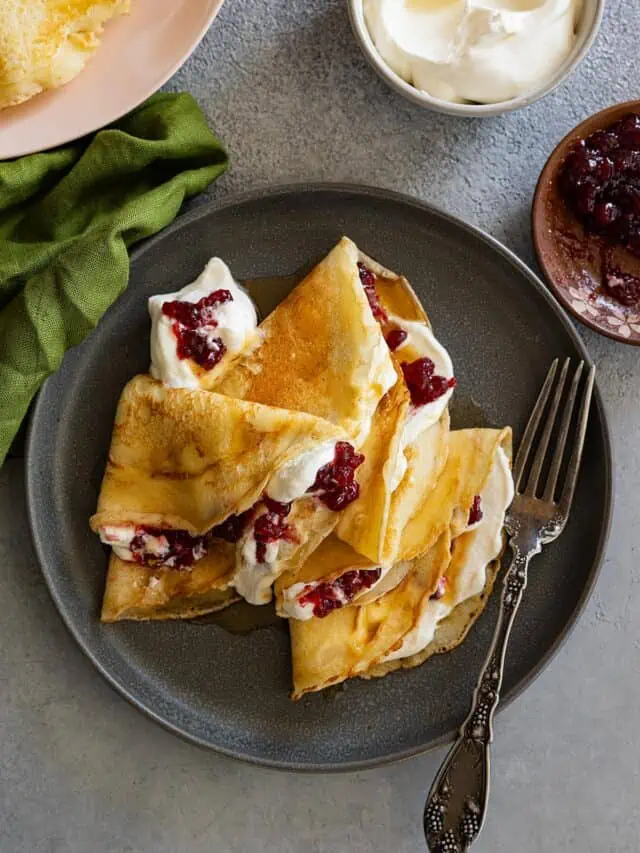 Overhead view of Swedish pancakes on a dark plate. They have whipped cream and lingonberries inside. Fork and green napkin to the side.