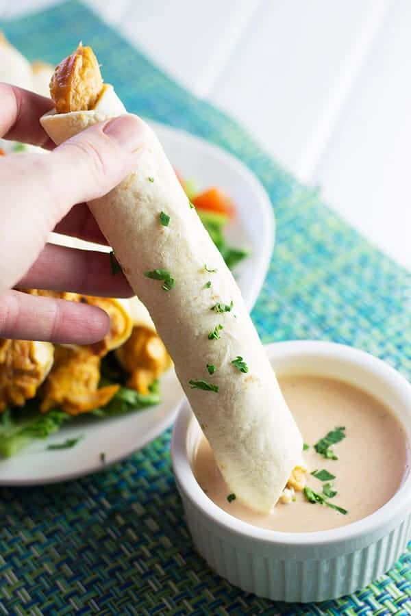closeup: a hand dipping my baked chicken taquitos into a ramekin with dipping sauce