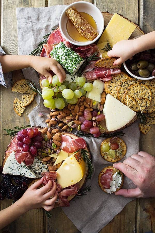 overhead: several hands reaching for an easy cheese board made with fruit, nuts, meat, olives, and crackers