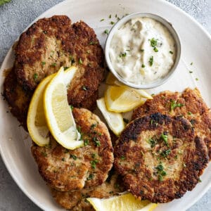 Overhead view of tuna patties on a white platter with tarter sauce and lemon wedges.