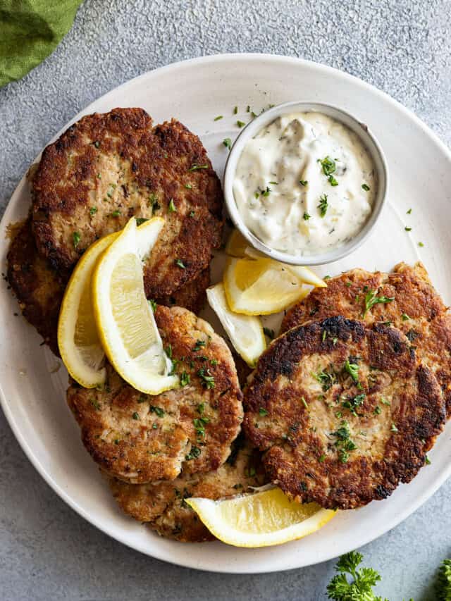 Overhead view of tuna patties on a white platter with tarter sauce and lemon wedges.