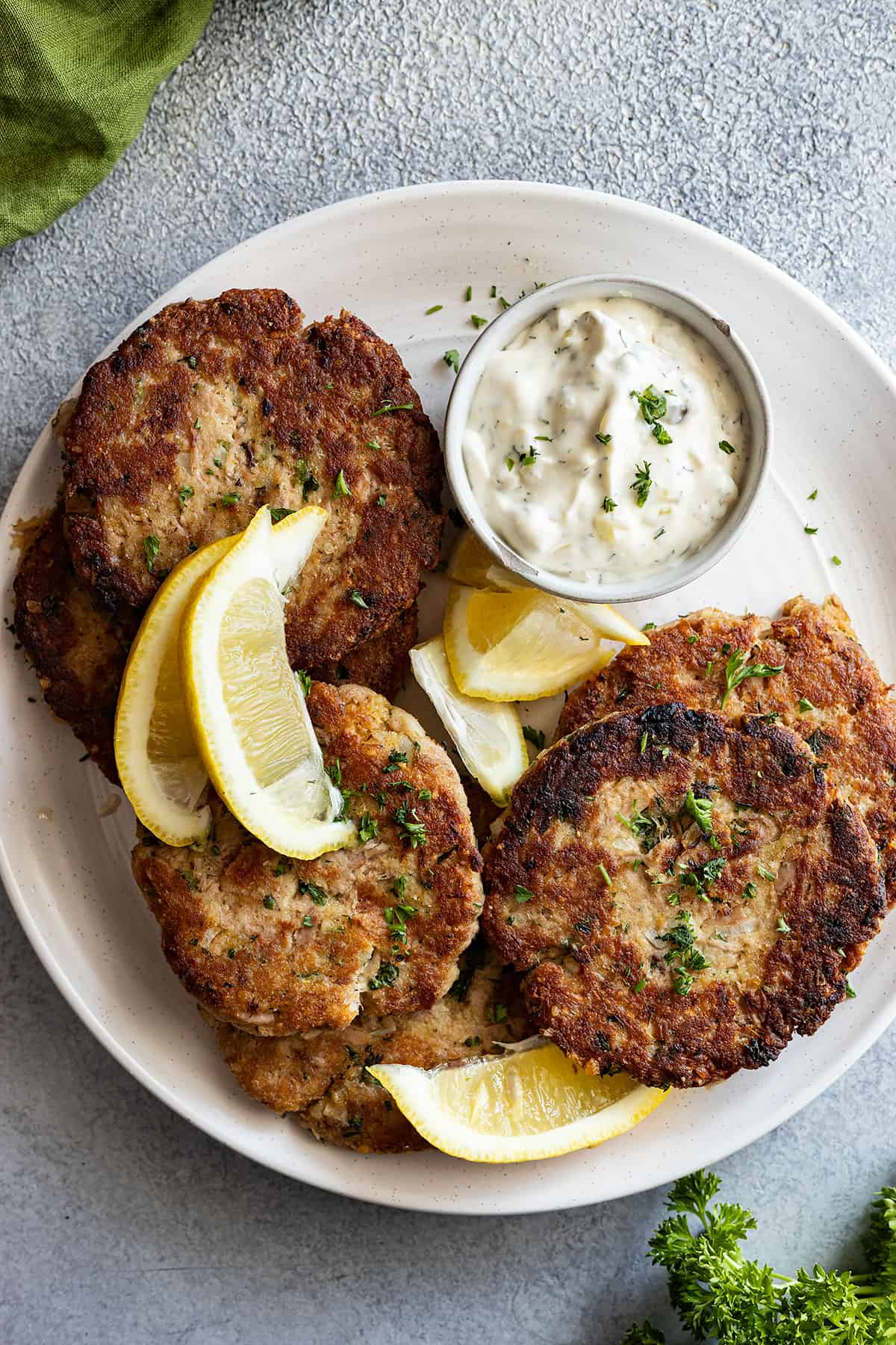 Overhead view of tuna patties on a white platter with tarter sauce and lemon wedges. 