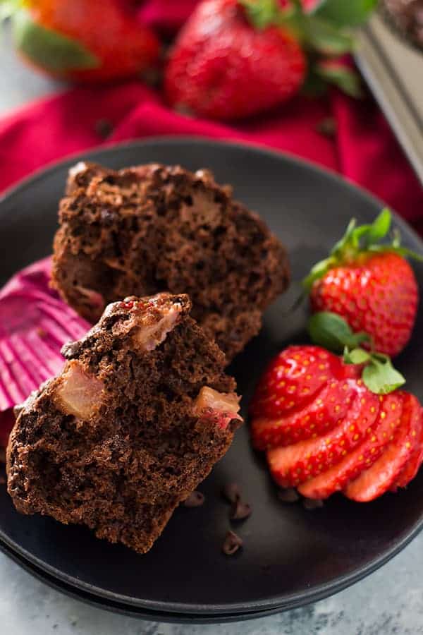 a double chocolate chip muffin sliced in half on a black plate with fresh strawberries and a paper liner to the side