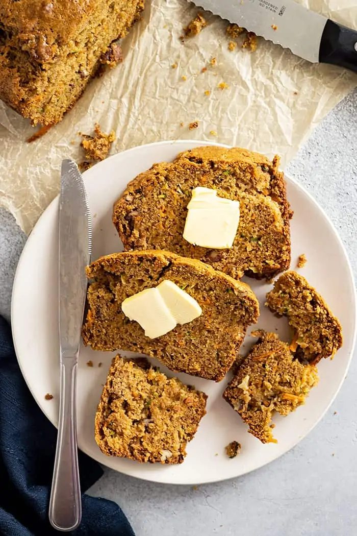 overhead: slices of zucchini carrot bread on a plate topped with pats of butter with a knife and a napkin off to the side