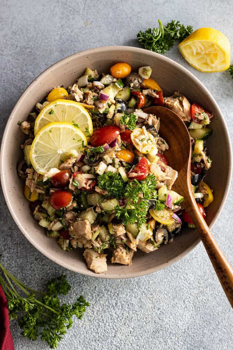 Overhead view of tuna salad in a large bowl with wooden spoon in it. Garnished with fresh herbs and lemon slices.