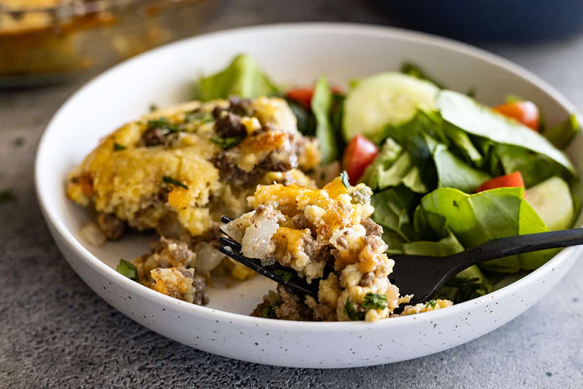 A serving of impossible cheeseburger pie in a white shallow bowl with a fork taking a bite out. 
