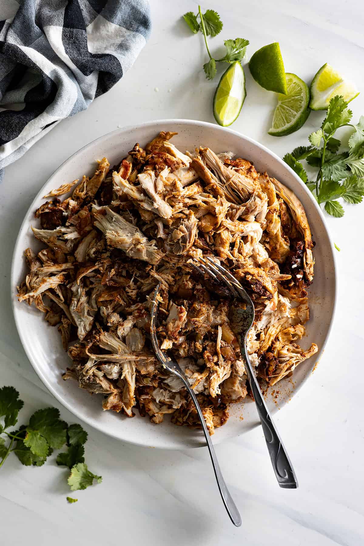 Overhead view of chicken taco filling in a white bowl with forks in it and cilantro and limes off to the side. 