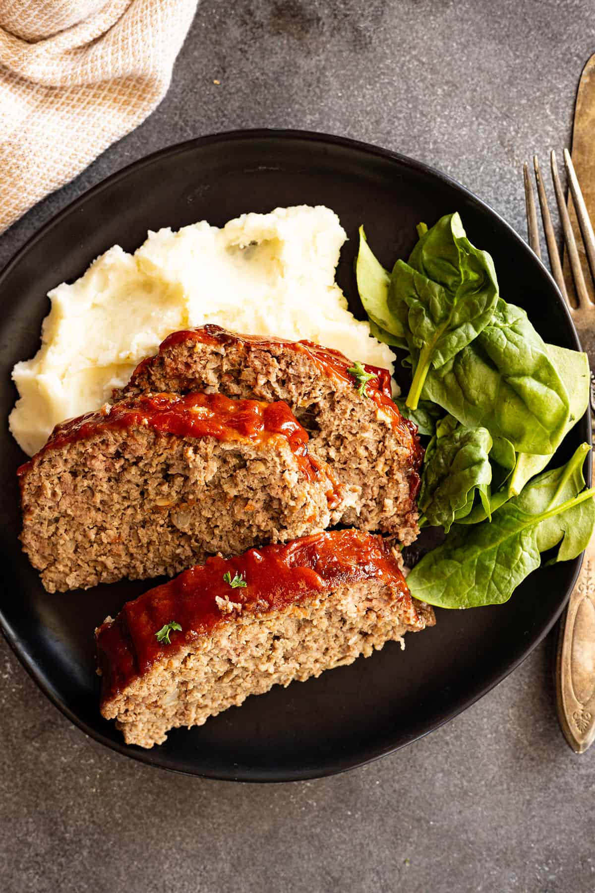 Meatloaf slices on a black plate with mashed potatoes and salad. 