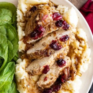 Cut up slices of baked cranberry chicken on a bed of fluffy mashed potatoes with a spinach salad to the side.