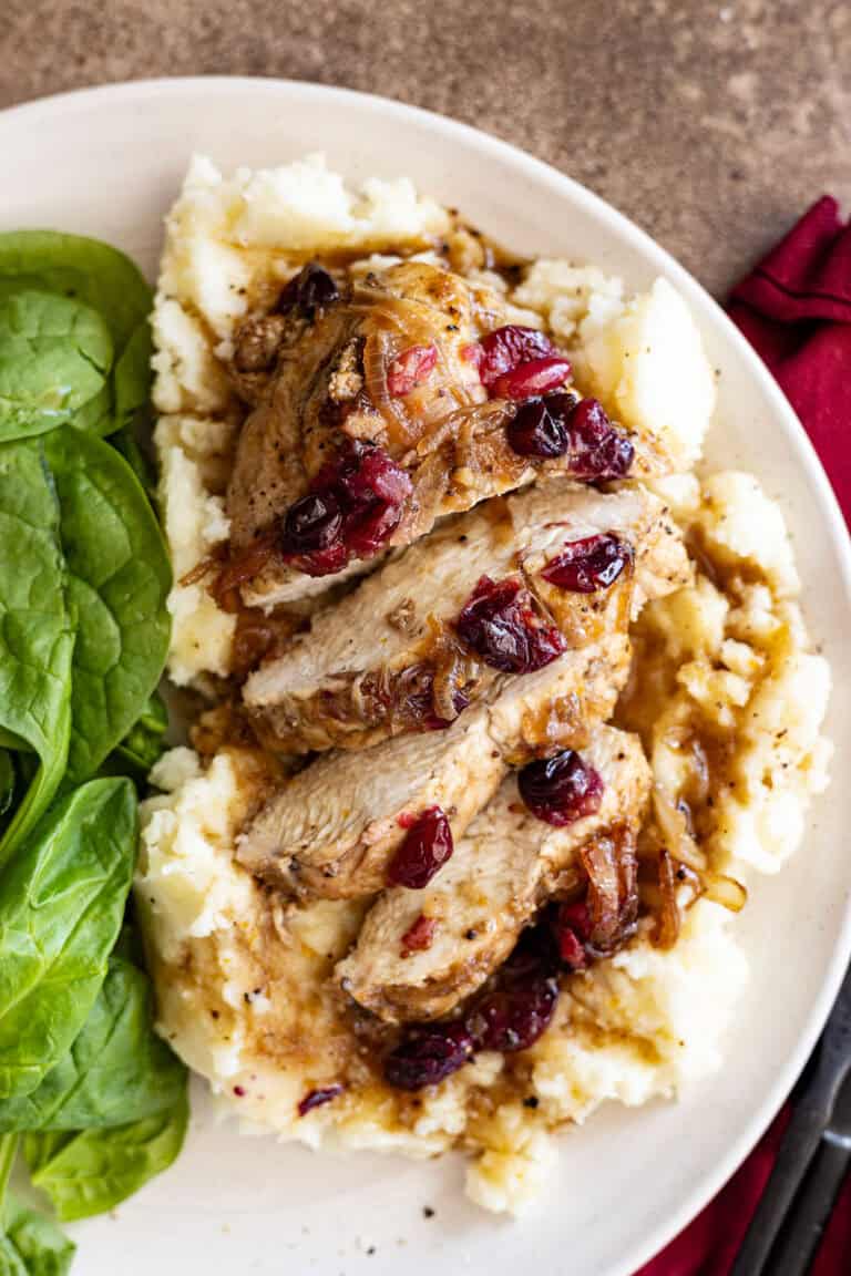 Cut up slices of baked cranberry chicken on a bed of fluffy mashed potatoes with a spinach salad to the side.
