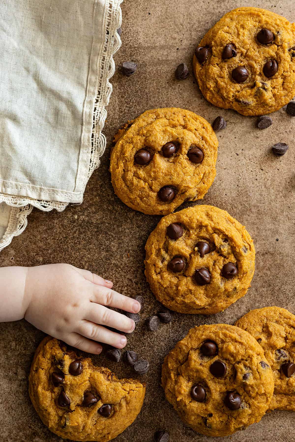 Cookies scattered along with chocolate chips and a cute baby hand reaching for a cookie. 