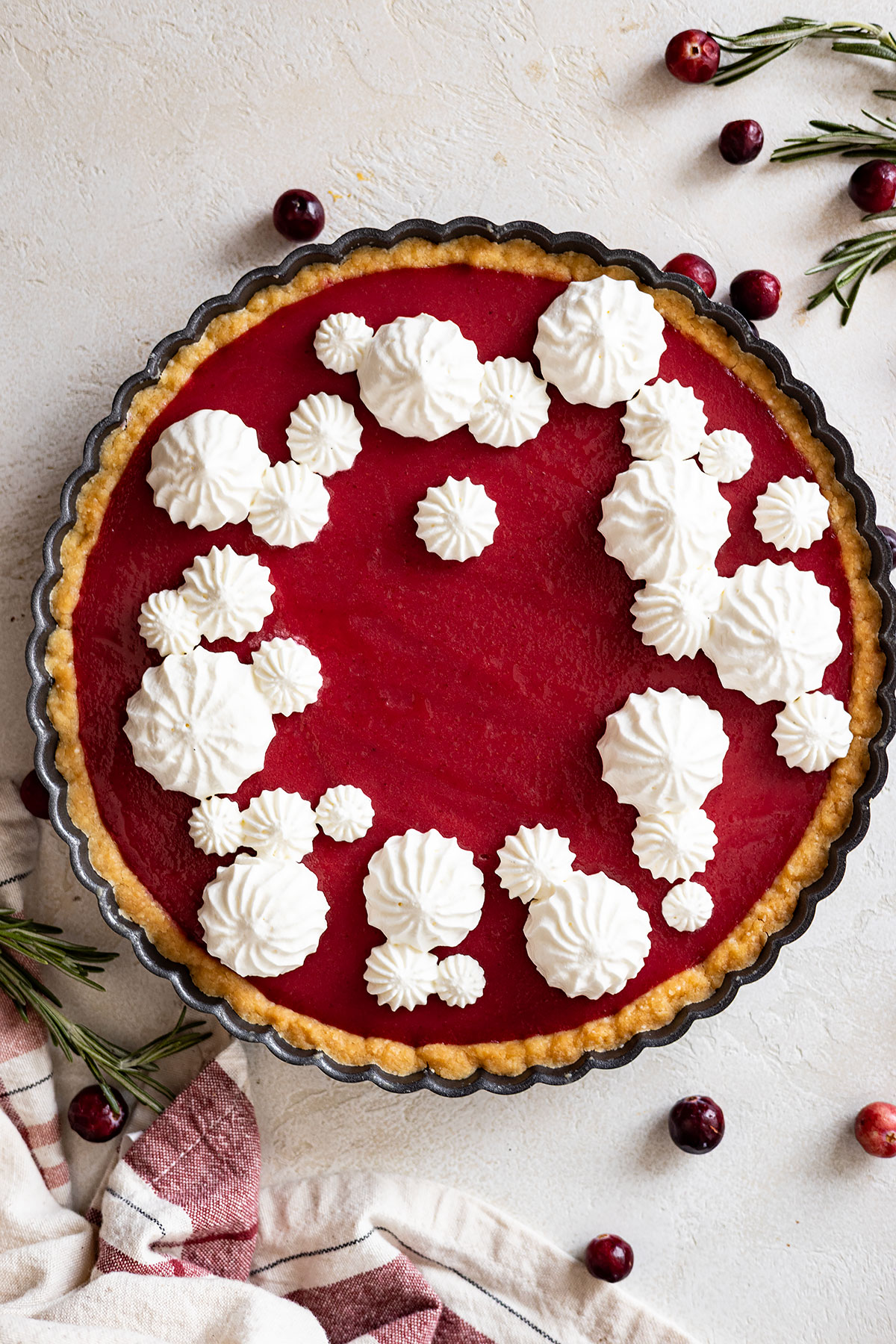 overhead view of cranberry curd tart decorated with whipped cream. rosemary and cranberries scattered around. 