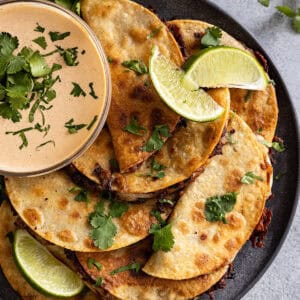 Overhead view of crispy black bean tacos stacked on a plate with a bowl of sauce on the side.