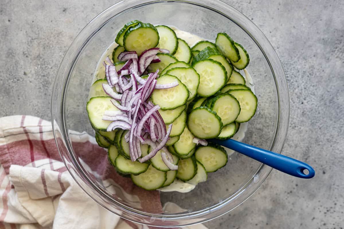 Overhead view of cucumbers and onion added to the bowl with the dressing.